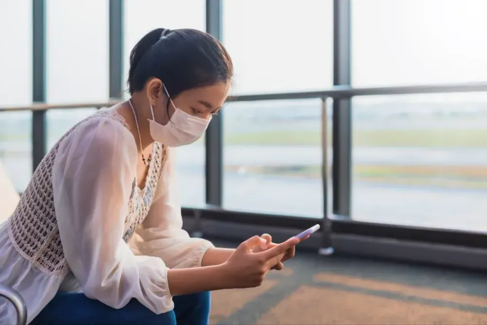 Woman sitting in airport terminal, wearing a face mask and using her smartphone